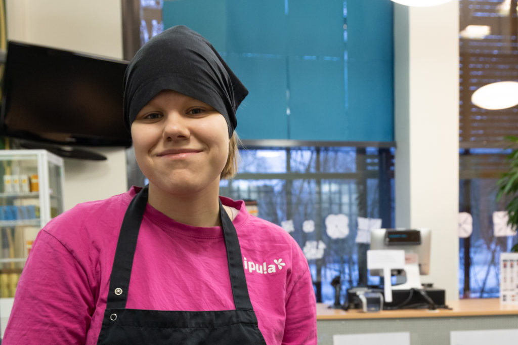 Lähikuva hymyilevästä nuoresta naisesta kahvilatyöntekijän asussa.