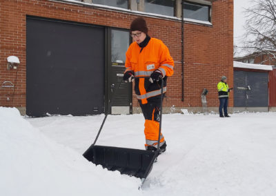 Opiskelija työvaatteissa työntää lunta lumikolalla ison tiilirakennuksen edessä.