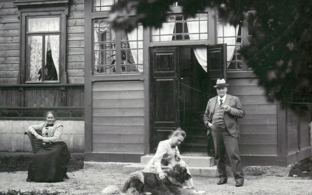 Vanhassa mustavalkoisessa kuvassa talon edessä Wilhelmiina Kiipula istuu tuolilla, Alma Kiipula silittää koiraa ja Kustaa Kiipula seisoo.