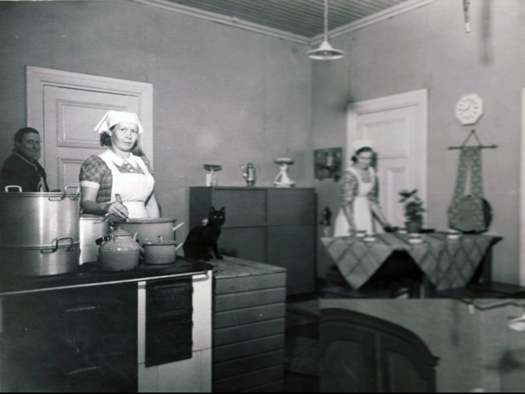 Vanha mustavalkoinen valokuva, jossa keittiön emäntä hämmentää kattilaa tuvassa.