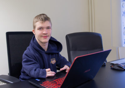Opiskelija hymyilee kannettavan tietokoneen takaa.