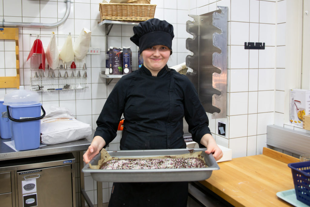 Mustissa kokkivaatteissa oleva opiskelija Venla pitää kädessään pellillistä valmiita mokkapaloja opetuskeittiössä. Hän hymyilee leveästi.