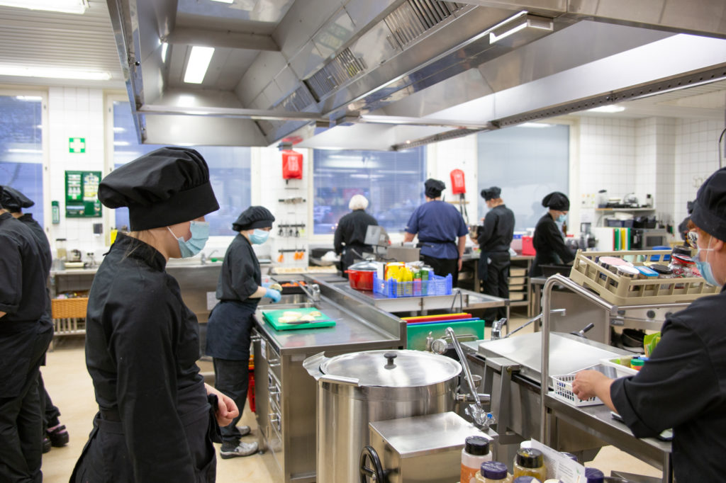 Opetuskeittiö, jossa näkyy kuusi opiskeiljaa mustissa keittiövaatteissa.