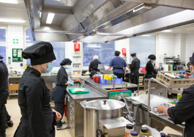 Opetuskeittiö, jossa näkyy kuusi opiskeiljaa keittiövaatteissa.