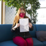 Maski päässä oleva Mea Vartiainen isuu sinisellä sohvalla ja pitää käsissään Taitaja-finalisti-kunniakirjaa.