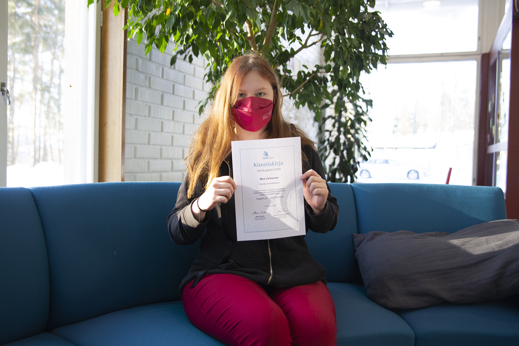 Maski päässä oleva Mea Vartiainen isuu sinisellä sohvalla ja pitää käsissään Taitaja-finalisti-kunniakirjaa.
