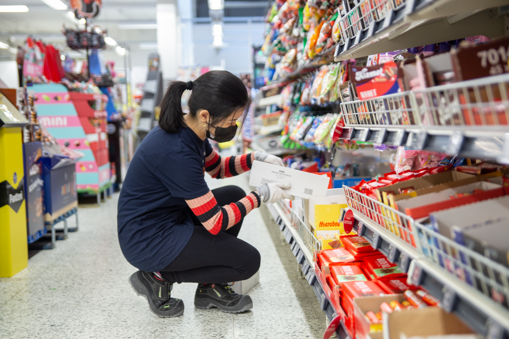 Merkonomiopiskelija hyllyttää karkkipatukoita supermarketin karkkiosastolla.