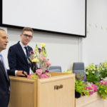 Opiskelija seisoo puhujakorokkeen takana auditoriossa. Rehtori Jukka Kujala seisoo vierellä.