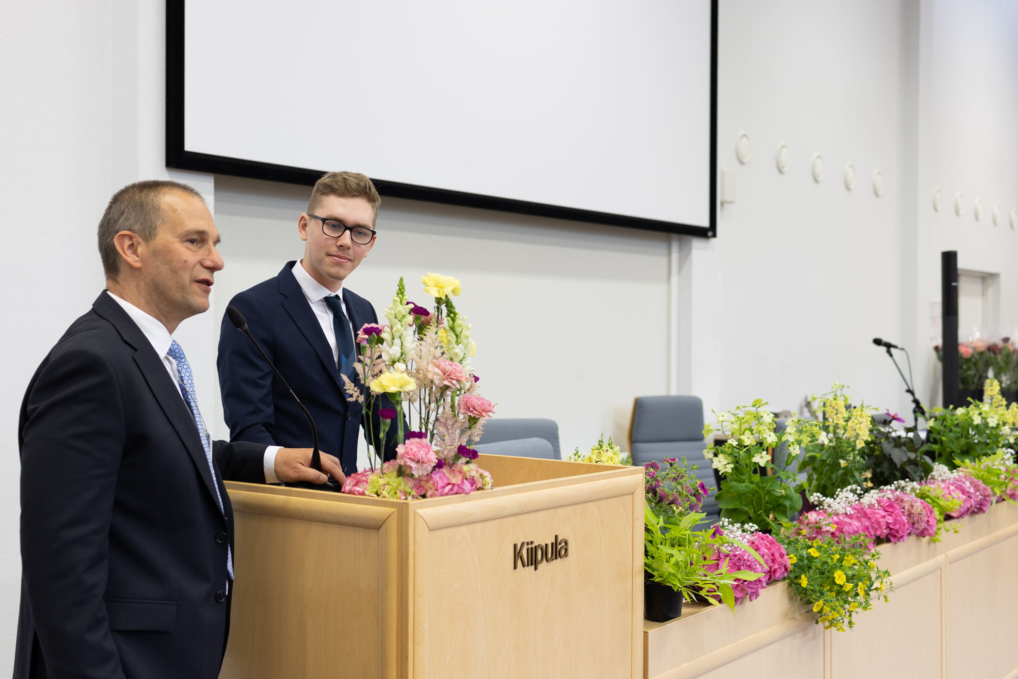 Opiskelija seisoo puhujakorokkeen takana auditoriossa. Rehtori Jukka Kujala seisoo vierellä.