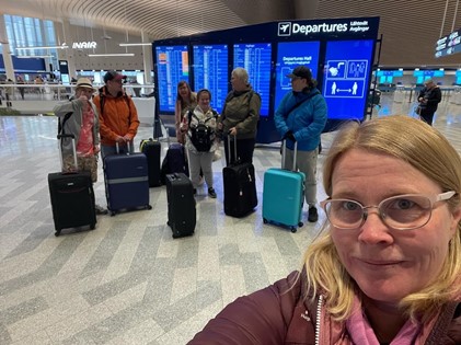 Lentoasemalla otettu selfie jossa taustalla näkyy joukko matkustajia laukkuineen.