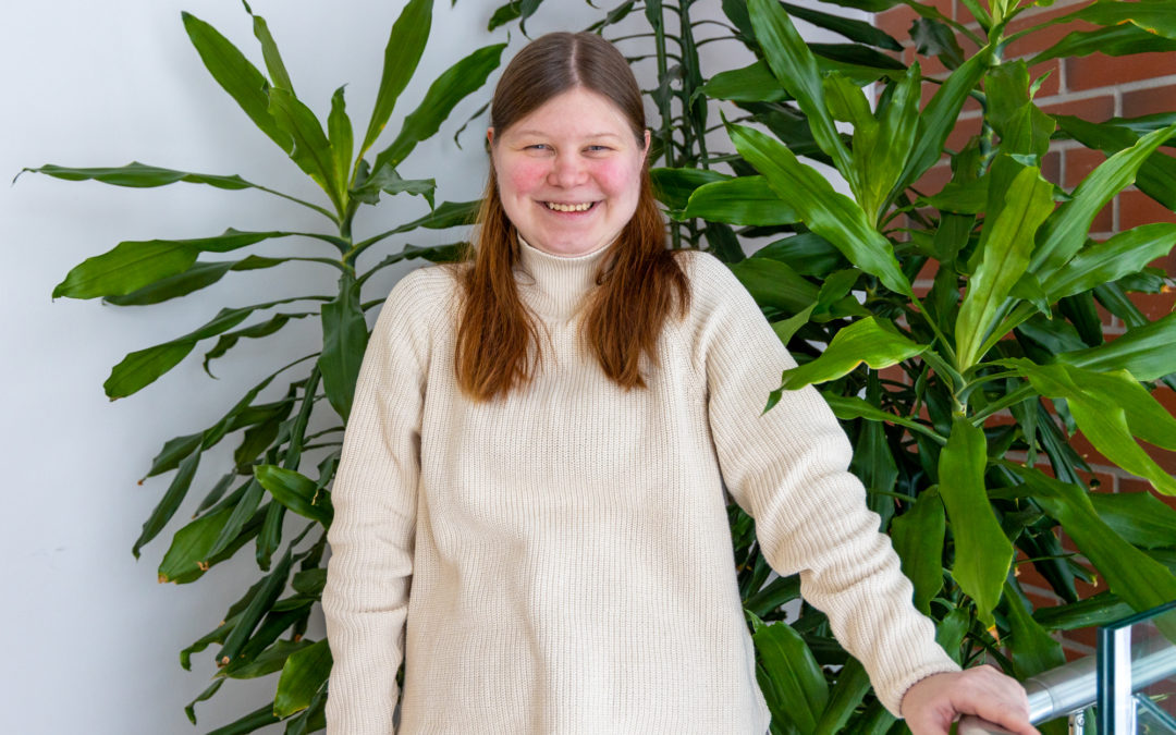 Valkoiseen villapaitaan pukeutunut opiskelija Merja hymyilee kameralle. Taustalla viherkasveja.