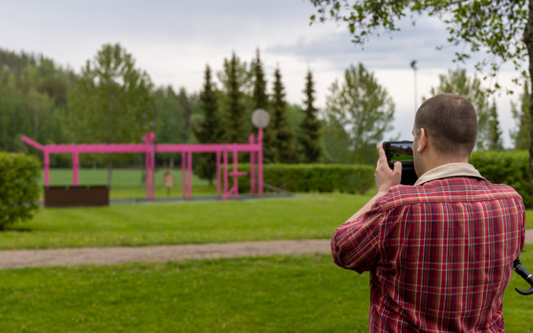 Mies kuvaa kännykällä puistoa, jossa taustalla näkyy magenta cross training -teline.