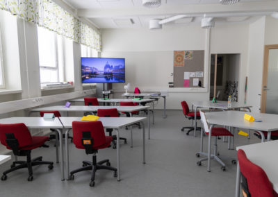 Tyhjä luokkahuone, jossa on pöydät ja punaiset työtuolit. Luokassa on iso taulutelevisio.