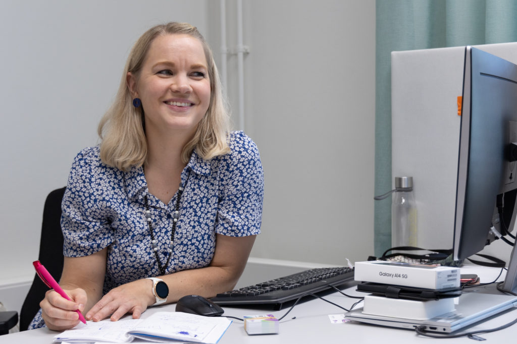 Koulutuspäällikkö Noora Häyhä istuu työpöydän takana ja hymyilee.